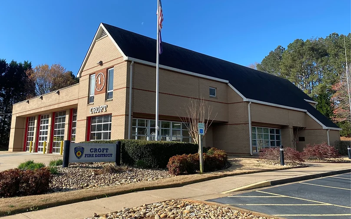 A one-story brick fire station with a steep black roof, bearing a sign that reads "CROFT." The building has a large garage with red doors and a flagpole with an American flag in front. A sign by the entrance reads "CROFT FIRE DISTRICT." Trees are in the background.
