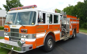 A bright orange and white fire truck from the Roebuck Fire Department is parked on a paved surface with a brick building and trees in the background.