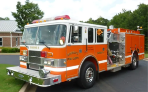 A bright orange and white fire truck labeled "Roebuck Fire Dept, Engine 2" is parked on a street near a brick building. The truck has visible equipment compartments and a "Slow" sign mounted on the side. Lush green trees are in the background.