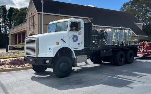A white fire department truck is parked outside a brick fire station. The flatbed of the truck carries a large water tank and equipment. The station building has a sign above the entrance.