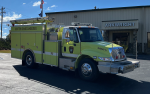 A green fire truck parked outside a building labeled "Arkwright." The truck has "South Spartanburg" written on its side. The sky is clear and sunny.