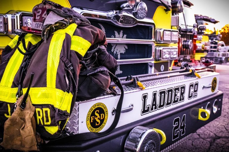A firefighter's gear is neatly placed on the bumper of a firetruck labeled "Ladder Co 24." The truck is black and yellow, with a helmet and jacket prominently displayed. The background shows other firetrucks and equipment under a dusky sky.