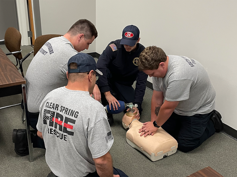Four emergency responders are practicing CPR on manikins in a classroom. Two are giving compressions, while another observes, and the fourth is using a medical device. They're wearing uniforms with "Clear Spring Fire & Rescue" logos.