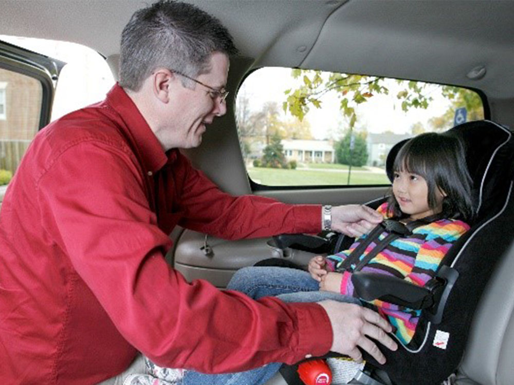 An adult helps a young child in a colorful striped sweater buckle into a car seat inside a vehicle. The child is smiling, and the car is parked with a view of trees and houses visible through the window.