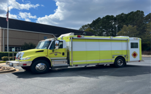 A bright yellow fire truck parked on a paved surface near a building with an American flag. The truck has "City of Dunwoody" visible on its side. Trees and a cloudy blue sky are in the background.
