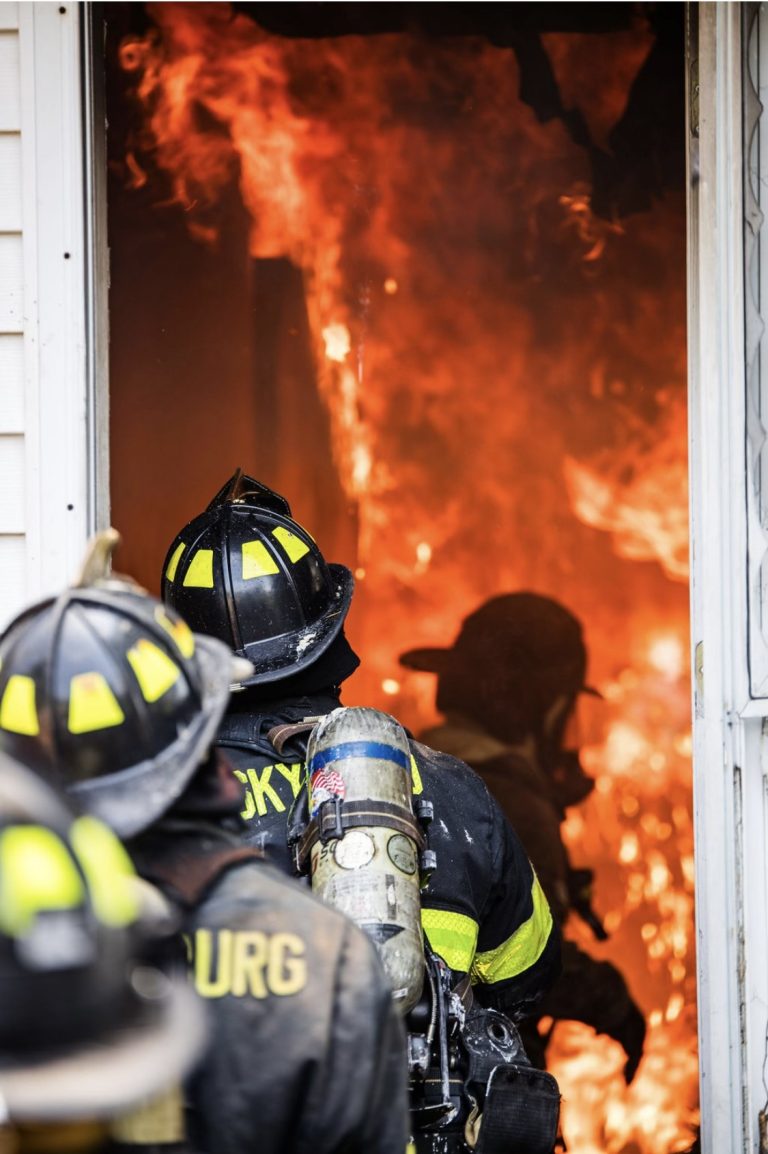 Firefighters, wearing protective gear and helmets, enter a building engulfed in flames. The scene shows intense fire inside, with firefighters focused on battling the blaze as they move through a doorway.