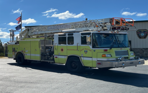 A yellow fire truck with a ladder parked in an outdoor area, under a clear blue sky. American flag and other flags are visible in the background, near a building with a badge insignia.