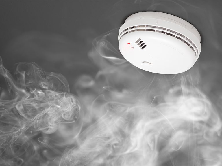 A smoke detector mounted on a ceiling surrounded by swirling smoke, with a red indicator light turned on.