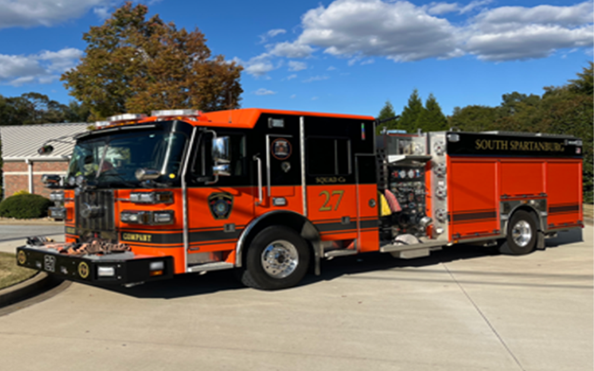A bright orange fire truck labeled "South Spartanburg" with the number 27 parked on a driveway. The vehicle is equipped with various emergency tools. Trees and a building are in the background under a blue sky with clouds.