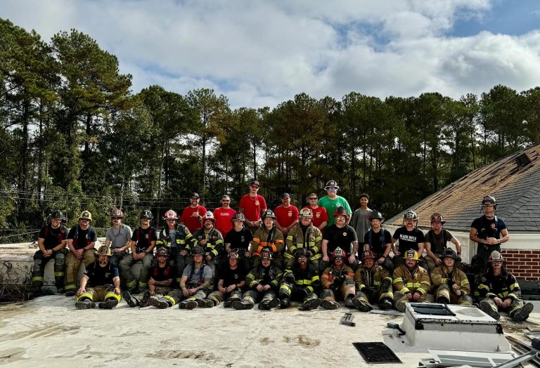 A group of firefighters in various uniforms and helmets pose together on a rooftop under a partly cloudy sky. Behind them, trees line the scene. Some wear red shirts, while others are in full firefighting gear. Equipment is visible on the roof.