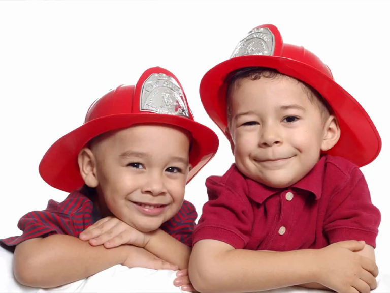 Two young children wearing red firefighter helmets smile at the camera. They are leaning on their arms, with a playful expression, against a white background.