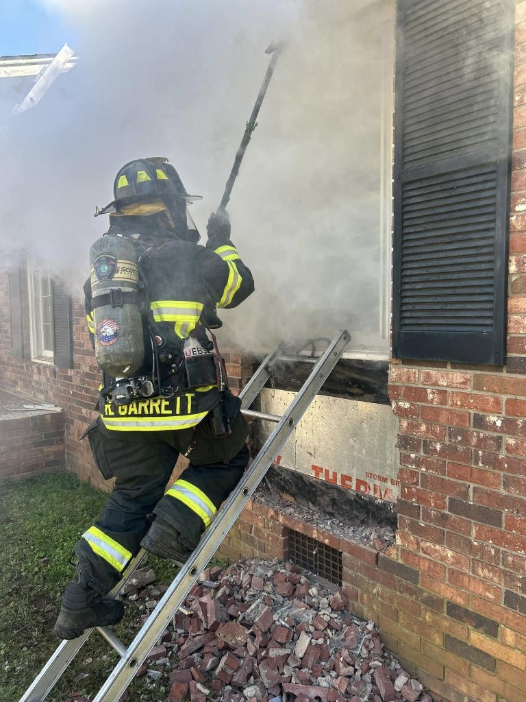 A firefighter in full gear stands on a ladder, breaking into a smoke-filled brick building through a window. There's rubble at the base of the wall, and the scene appears active with thick smoke surrounding the area.