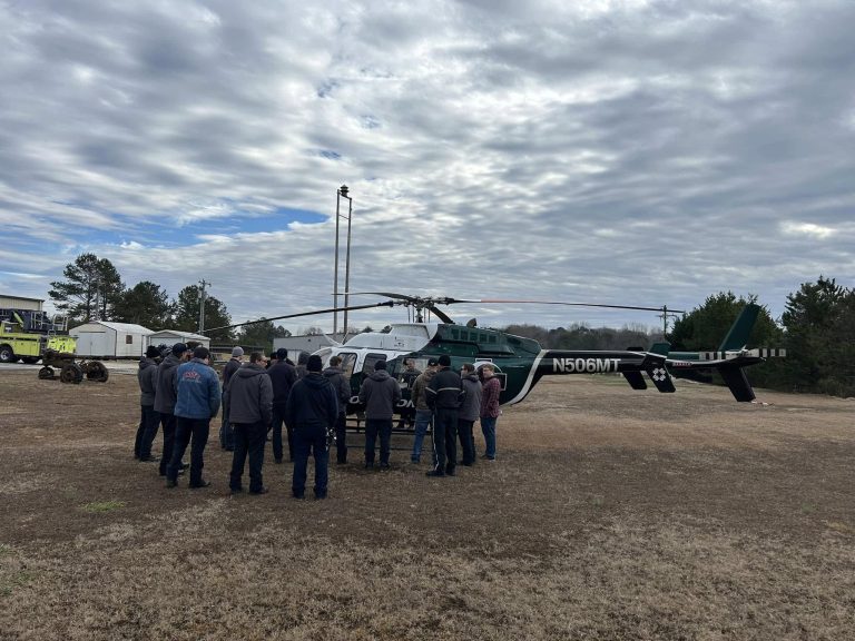 A group of people stand in a semicircle listening to a person in front of a helicopter marked N506MT. The sky is cloudy, and the scene is set on a grassy field with trees and a few vehicles in the background.
