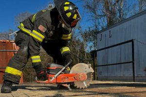 A firefighter in full gear and helmet is operating a power saw to cut through a wooden surface outdoors. There's a shipping container in the background, and the scene is set on a clear, sunny day.