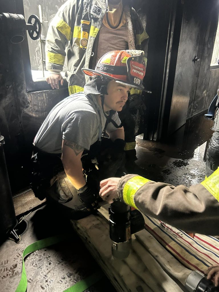 A firefighter in full gear is crouched down, holding equipment inside a dark, partially charred room. Smoke stains are visible on the walls. Another firefighter is partially visible, wearing a reflective jacket.