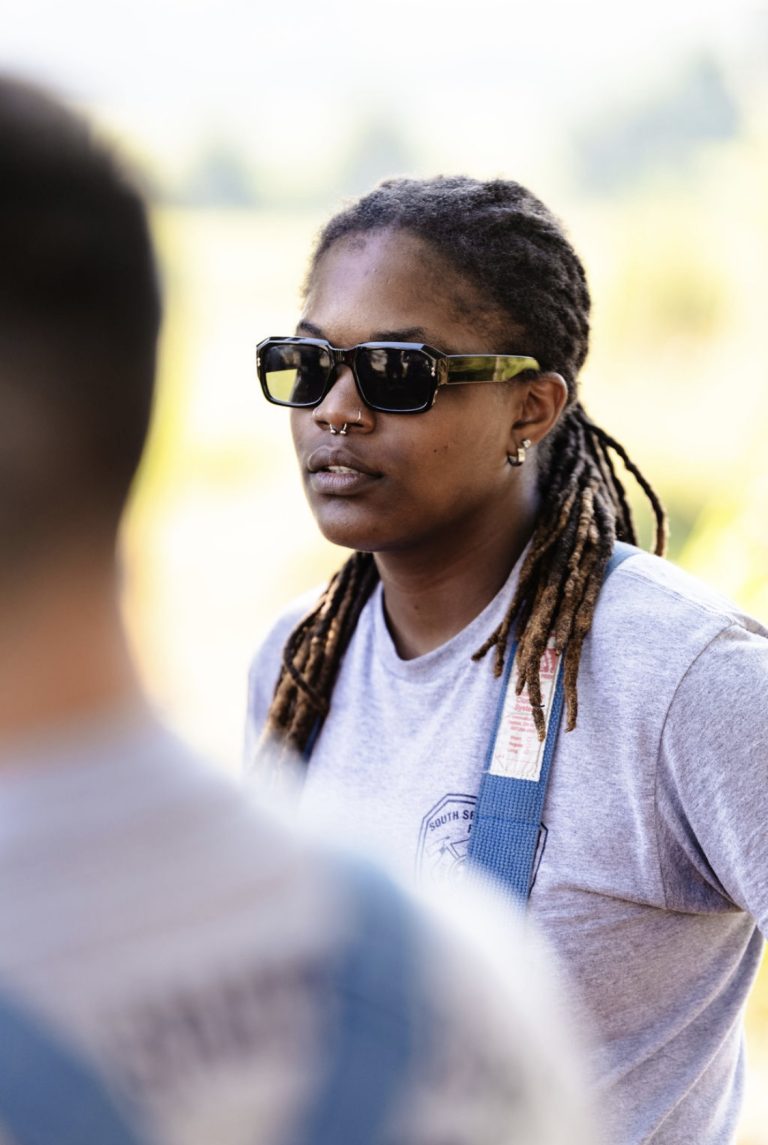 Person with sunglasses and long braided hair wearing a gray T-shirt stands outdoors. The background is blurred, with another person partially visible in the foreground, suggesting a sunny setting.