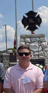 A person wearing sunglasses, a pink polo shirt, beige pants, and gray shoes stands in front of a stone memorial. They have a name tag on their shirt. The background includes flags, a wreath, and a sunny sky.