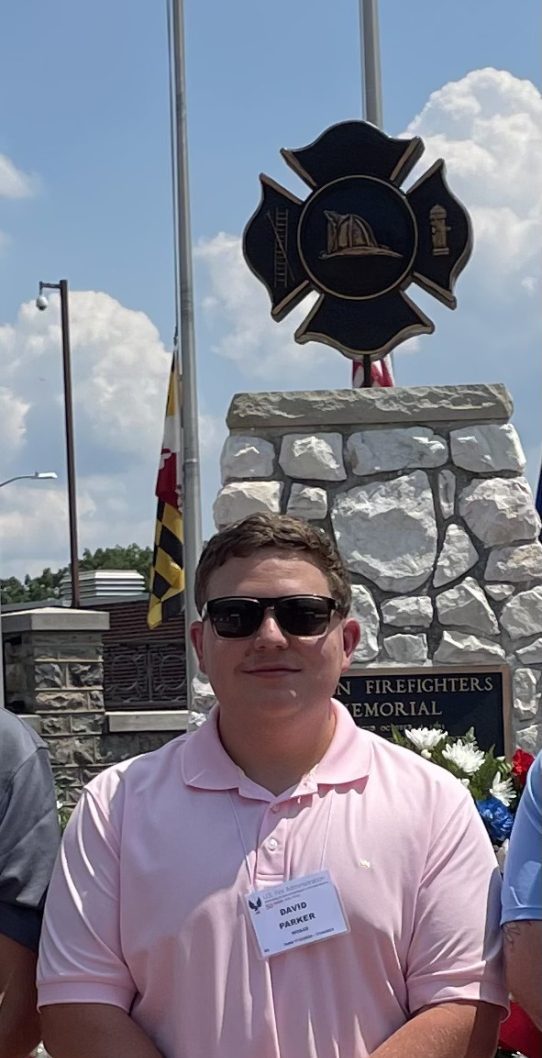 A person wearing sunglasses, a pink polo shirt, beige pants, and gray shoes stands in front of a stone memorial. They have a name tag on their shirt. The background includes flags, a wreath, and a sunny sky.
