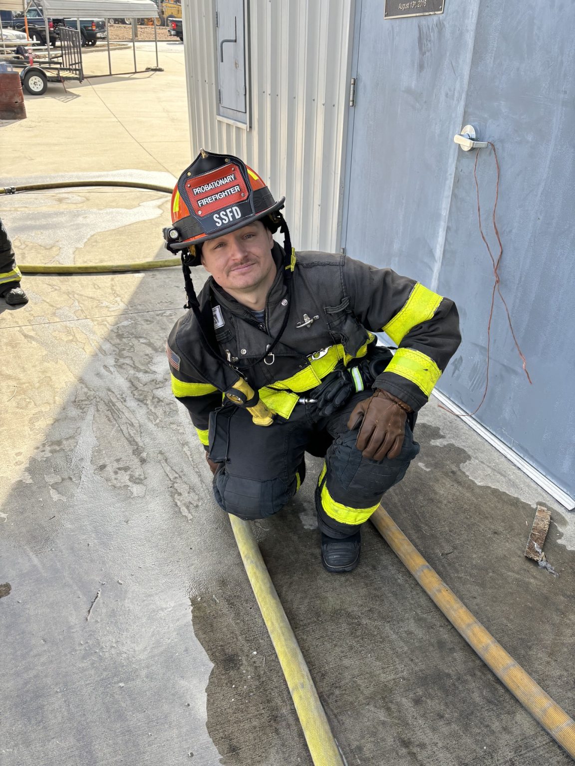 A firefighter in full gear, including a helmet, crouches next to a fire hose on a concrete surface. The building wall and door are visible behind, with several hoses laid out on the ground.