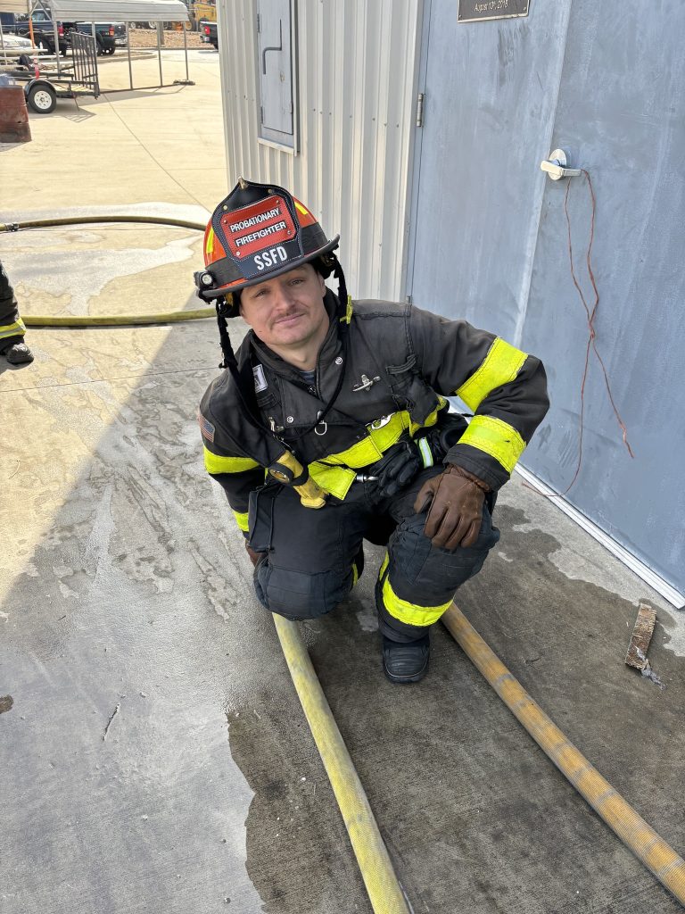 A firefighter in full gear, including a helmet, crouches next to a fire hose on a concrete surface. The building wall and door are visible behind, with several hoses laid out on the ground.