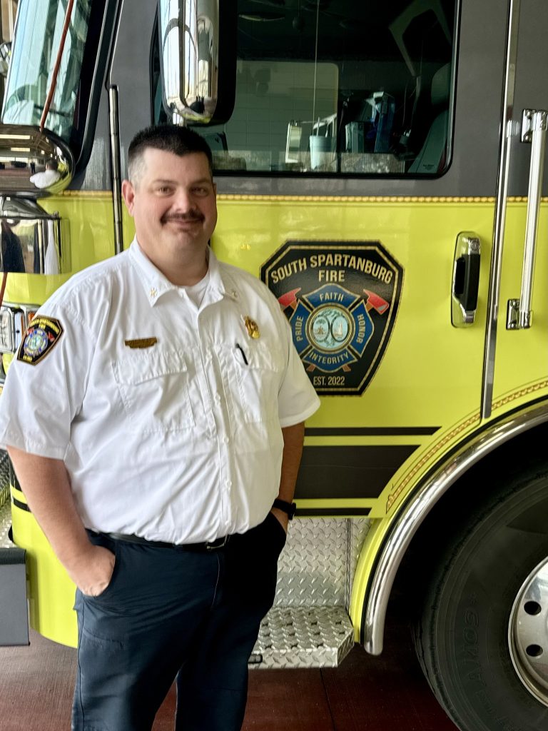 A person in a white uniform stands in front of a yellow fire truck with the South Spartanburg Fire Department emblem.