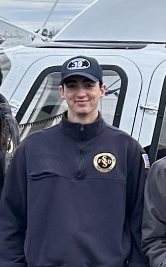 A person in a navy blue uniform and cap stands in front of a helicopter. The uniform features a logo and an American flag patch. The background shows parts of the helicopter and overcast skies.