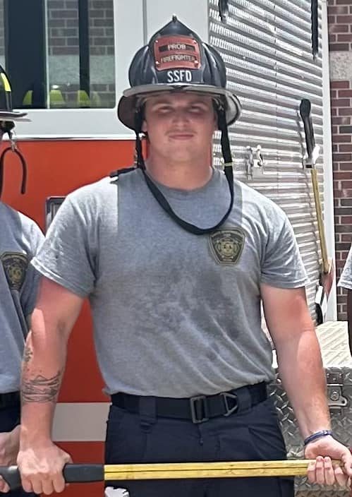 A firefighter stands in front of a fire truck, wearing a helmet and holding a tool. He is dressed in a gray T-shirt and dark pants, with visible tattoos on his arm. The setting is outdoors, next to a red fire truck with a metallic surface.