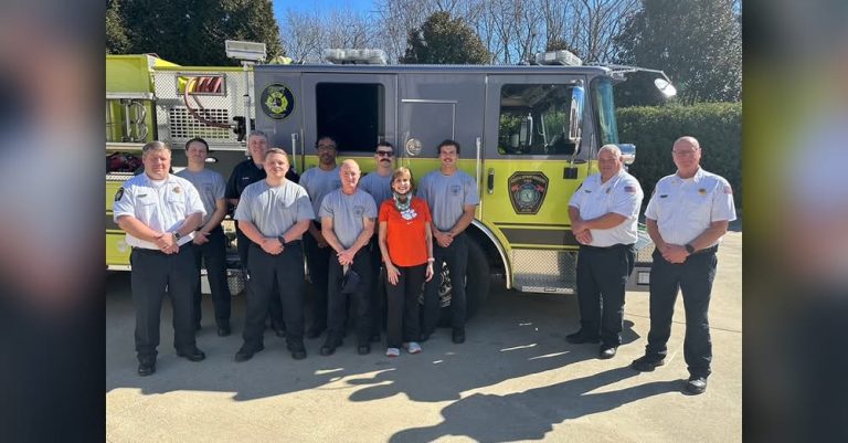 A group of firefighters and one woman stand smiling in front of a yellow fire truck on a sunny day, with trees and blue sky in the background.