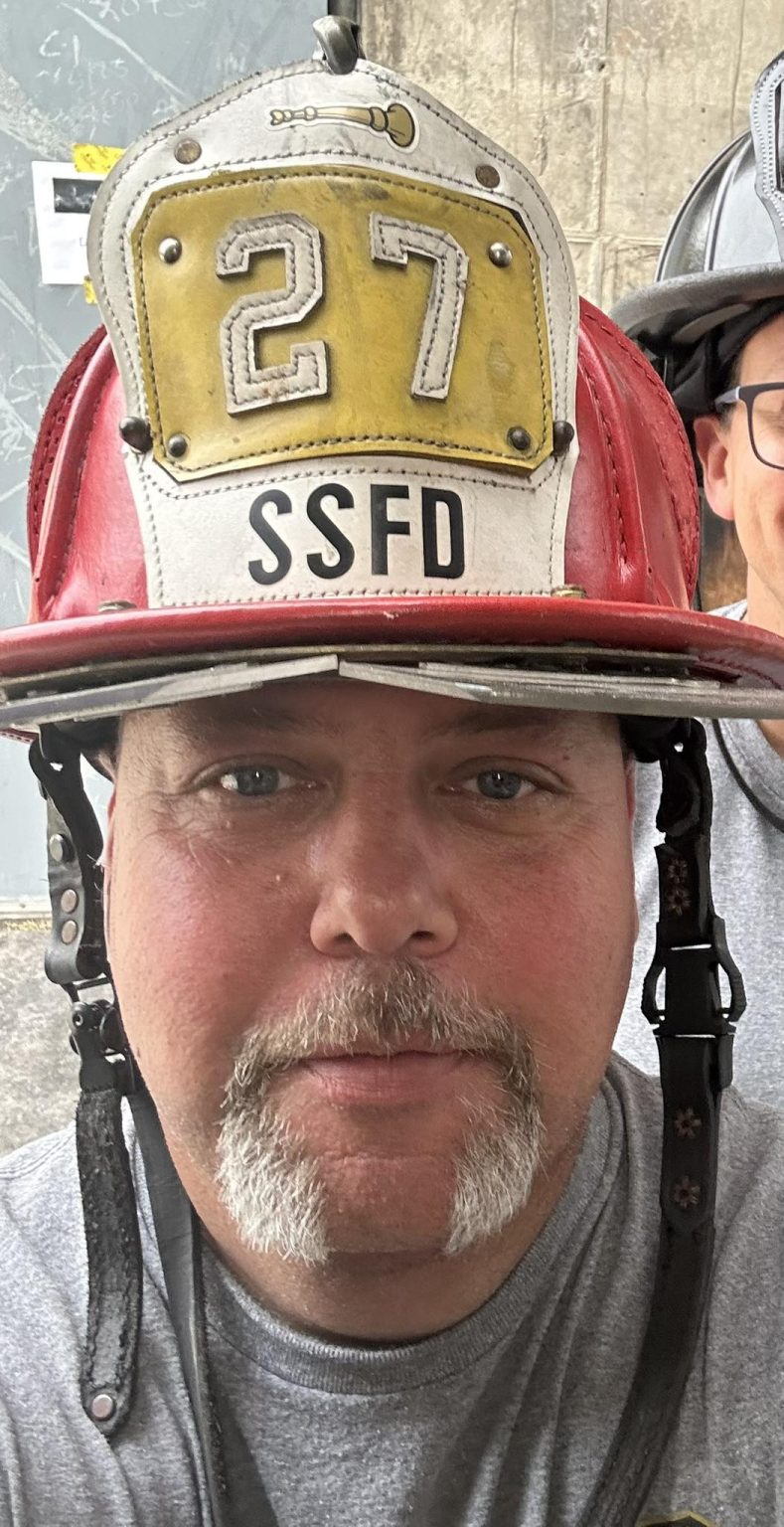 A firefighter with a gray mustache wears a red helmet labeled "27 SSFD." Part of another person's face is visible behind him. The background includes a gray wall and chalk markings.