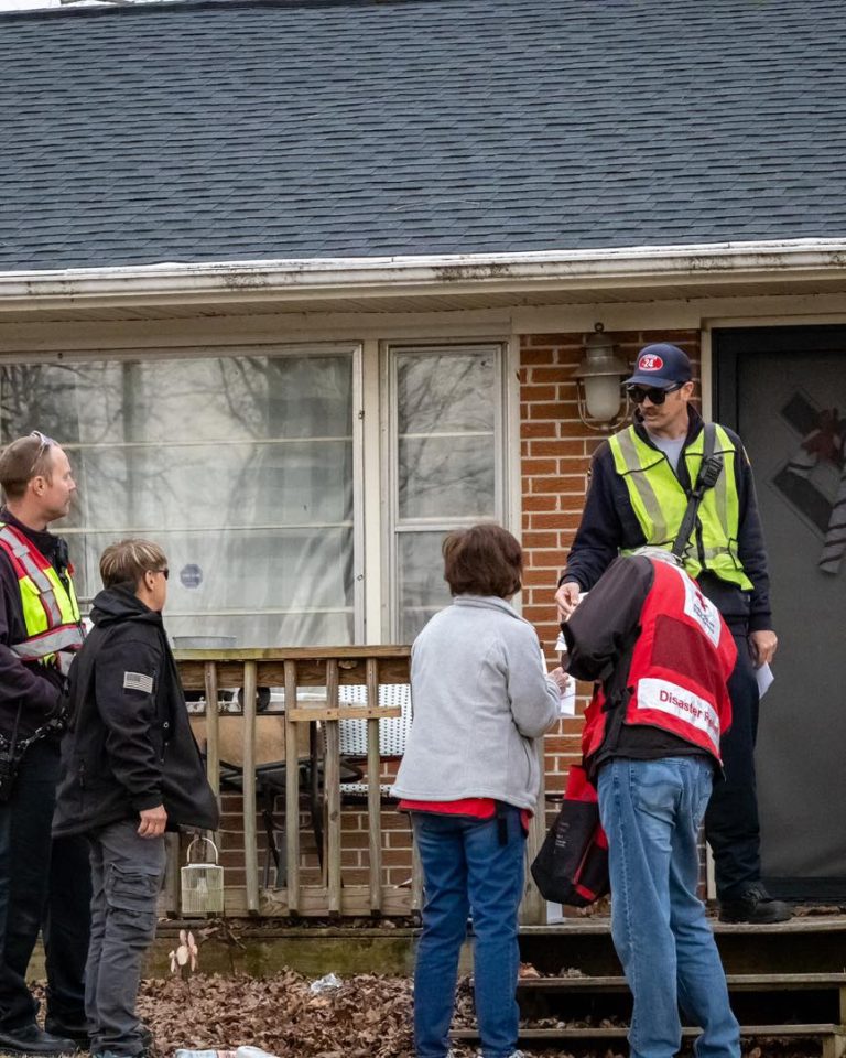 Four emergency responders in safety vests stand on the porch of a brick house, speaking with a resident. One responder hands out papers. The scene suggests community outreach or emergency response.