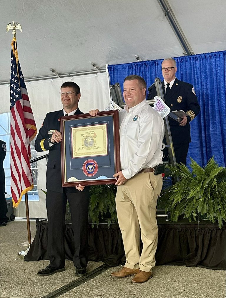 Two men stand smiling, holding a large framed certificate or award. One man wears a dark uniform, the other a white shirt and khakis. An official speaks at a podium behind them, with an American flag and blue curtains in the background.