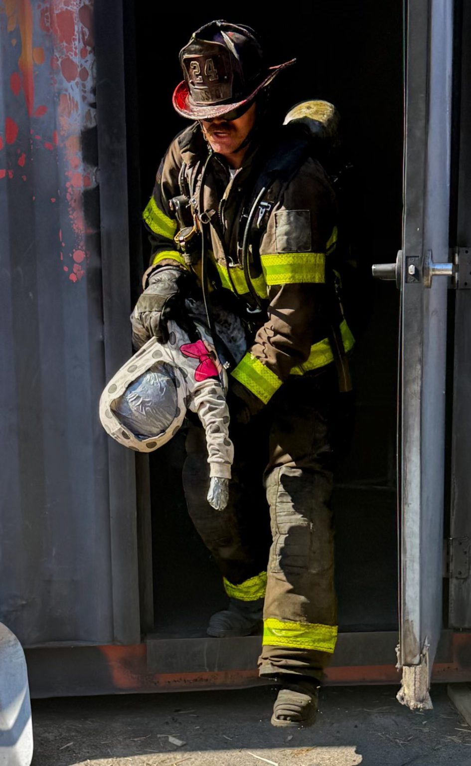 A firefighter in full gear carries a child-sized training dummy out of a dark, smoke-stained building during a rescue drill, sunlight highlighting their uniform’s reflective stripes.
