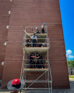 Several people wearing helmets and safety gear stand on three levels of scaffolding beside a tall brick building, appearing to participate in a training or construction exercise on a sunny day.
