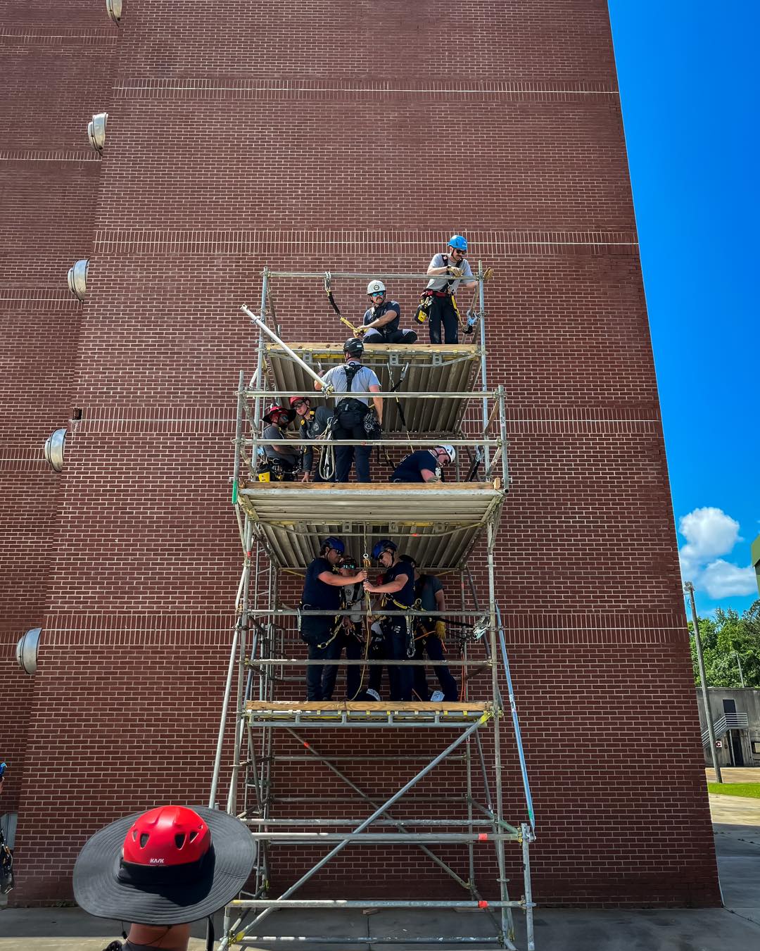 Several people wearing helmets and safety gear stand on three levels of scaffolding beside a tall brick building, appearing to participate in a training or construction exercise on a sunny day.