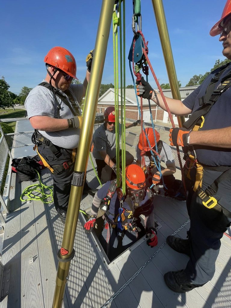 Four people wearing helmets and safety gear perform a rope rescue training exercise on a rooftop, using a tripod and harness system to lower a person into a confined space. The sky is clear and sunny.