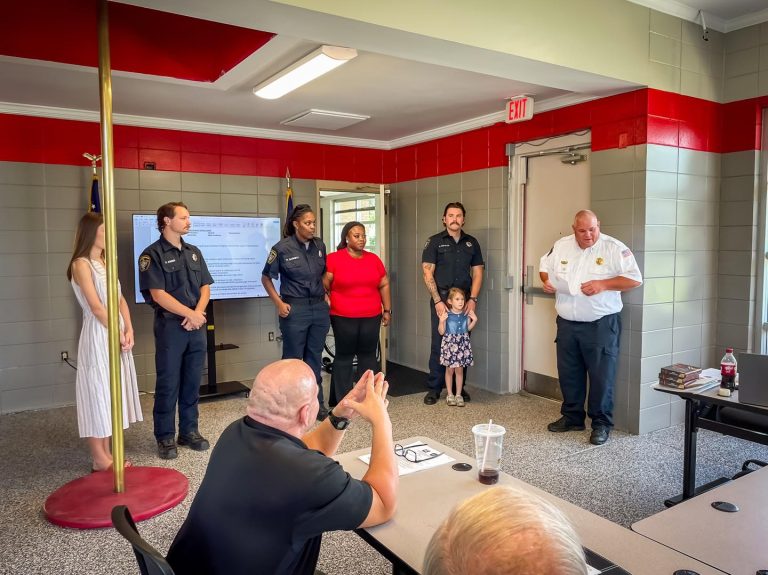 A group of firefighters and two civilians stand at the front of a room while a fire chief speaks; several people are seated, watching and applauding. A young girl stands holding hands with an adult.