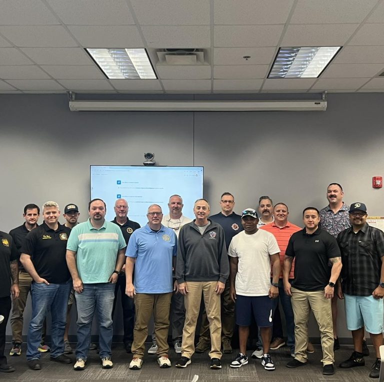 A group of sixteen men pose together in a classroom setting, standing in front of a screen and whiteboard. Most are dressed in casual or business casual attire, and all are smiling at the camera.