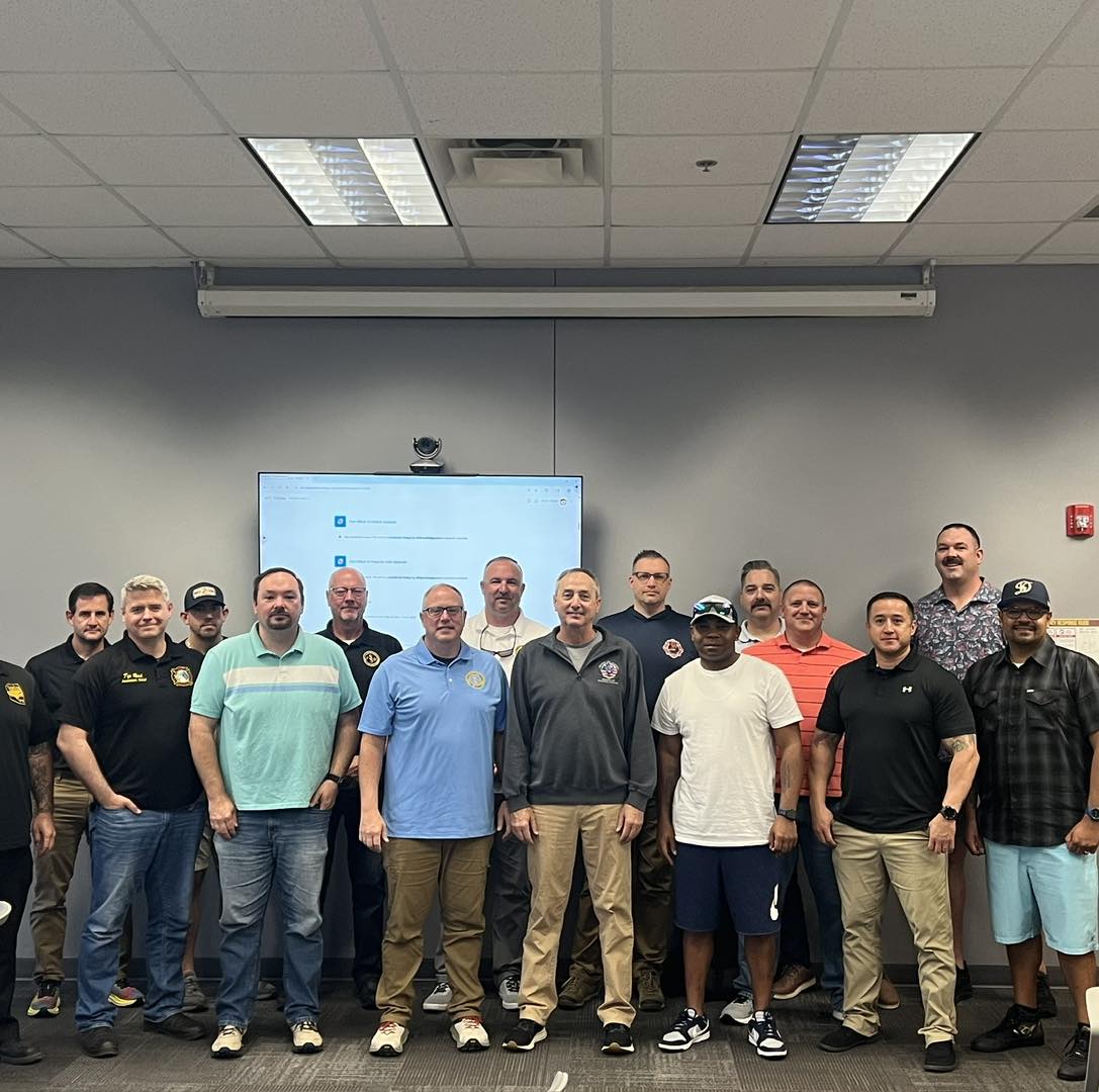 A group of sixteen men pose together in a classroom setting, standing in front of a screen and whiteboard. Most are dressed in casual or business casual attire, and all are smiling at the camera.