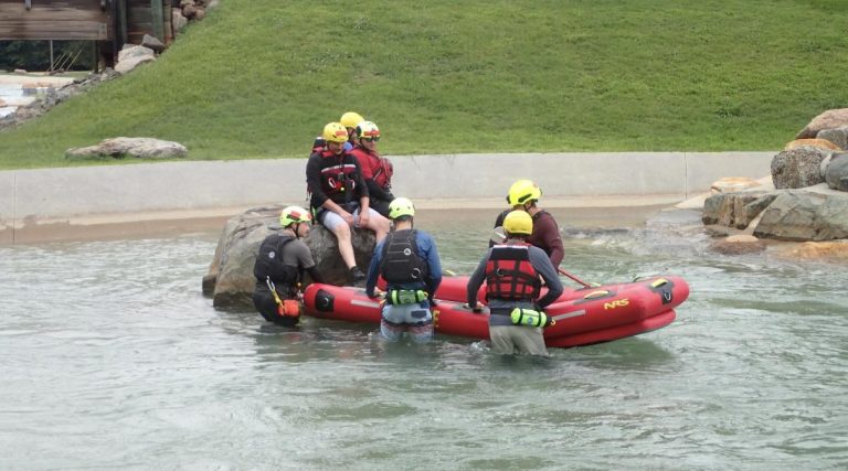 A group of people wearing helmets and life jackets assist an individual onto a red inflatable raft near a rocky edge in shallow water, with grassy banks in the background.