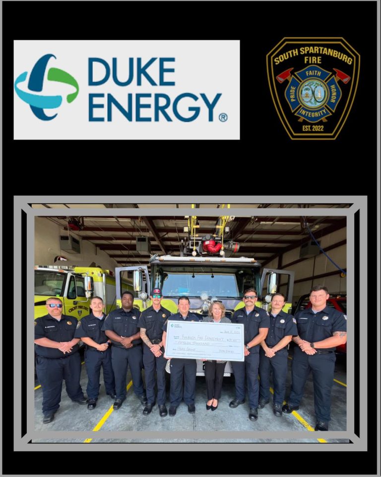 A group of firefighters and two civilians stand in front of a fire truck holding a large check. Above them are the Duke Energy logo and the South Spartanburg Fire Department badge.