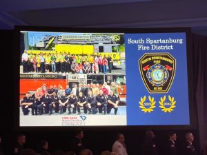 A presentation slide displays two group photos of firefighters in front of fire trucks, the South Spartanburg Fire District badge, and award emblems for 2023 and 2024. Text reads "South Spartanburg Fire District.