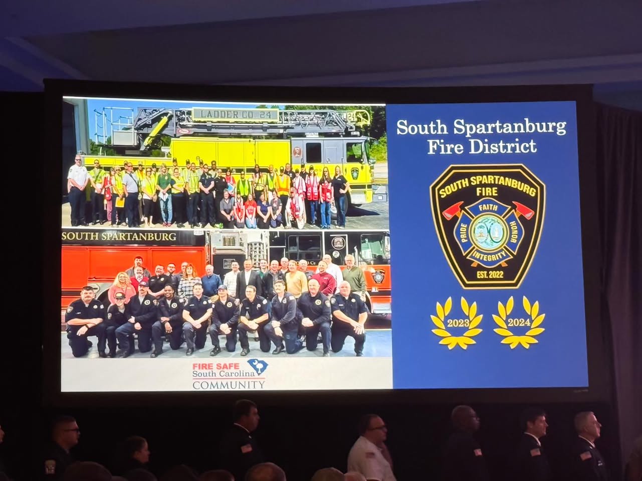 A presentation slide displays two group photos of firefighters in front of fire trucks, the South Spartanburg Fire District badge, and award emblems for 2023 and 2024. Text reads "South Spartanburg Fire District.