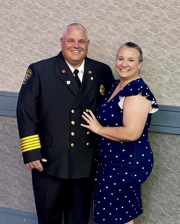 A man in a formal fire chief uniform stands smiling next to a woman in a blue polka dot dress, both posing happily against a neutral textured wall background.