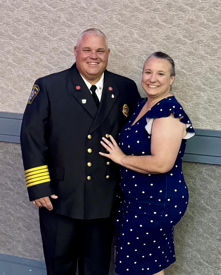 A man in a formal fire chief uniform stands smiling next to a woman in a blue polka dot dress, both posing happily against a neutral textured wall background.